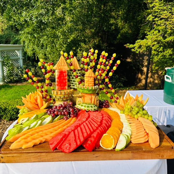 An elaborate fruit platter with carved melon castles and grape skewers, set outdoors.